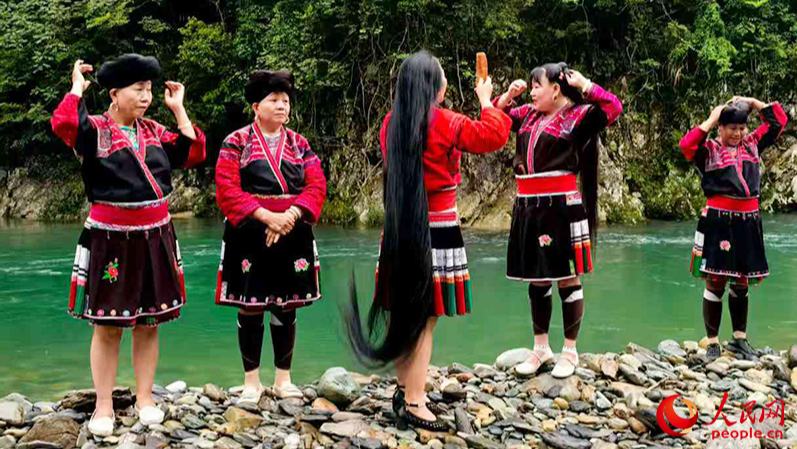 行進中國丨“長發(fā)”引客來雨后初晴，在廣西龍勝各族自治縣黃洛瑤寨的小河邊，幾名紅瑤婦女有說有笑地梳洗著如瀑布般傾瀉的長發(fā)。伴隨著流水聲，她們時不時齊聲合唱著《長發(fā)謠》，將長發(fā)盤于頭頂，再配上精美的發(fā)飾，形成“烏龍盤發(fā)”。